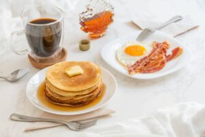 A cup of coffee, stack of pancakes topped with butter and syrup and a plate with a fried egg and 2 slices of bacon.