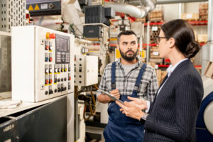 A man in a checkered shirt and blue overalls talks to a woman wearing a white shirt and pinstripe suit jacket. In the background is manufacturing equipment.