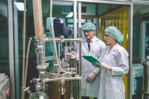 A man and a woman in white lab coats, aqua hairnets and safety goggles look at lab equipment.