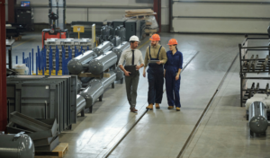 A man in a white shirt and white hard hat walks alongside a man and woman in blue overalls and orange hard hat, they are looking at a tablet whilst walking through a manufacturing plant.