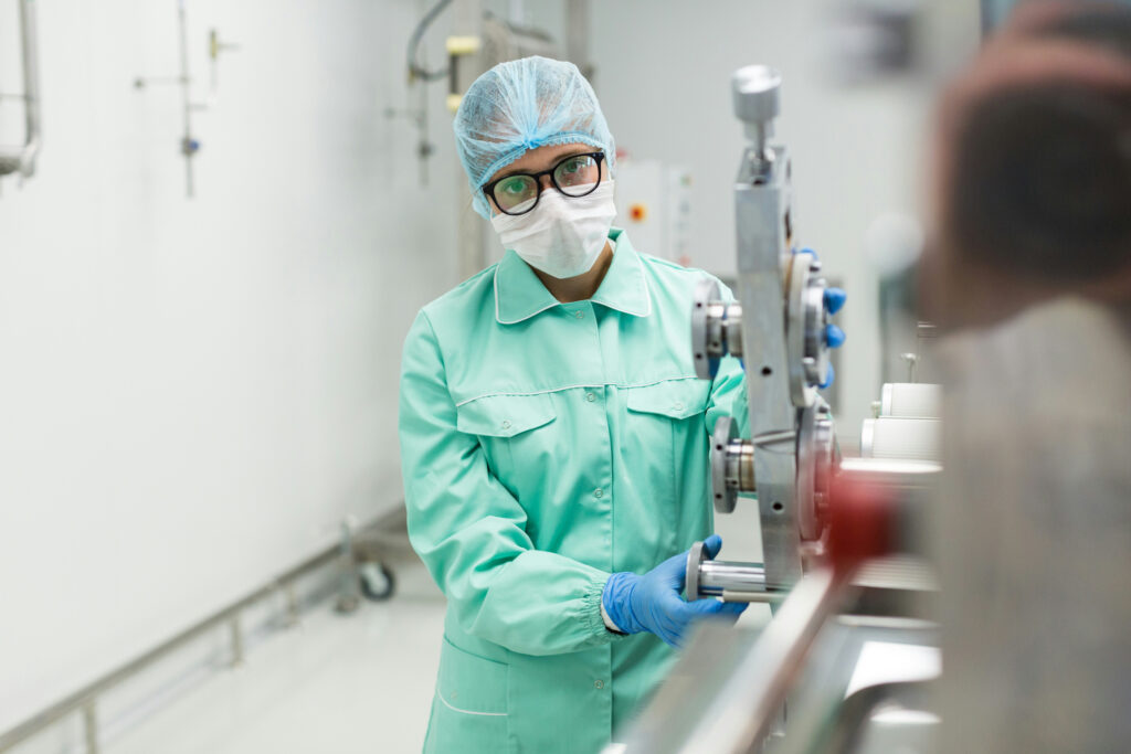 A woman wearing glasses, hairnet, blue PPE and latex gloves stands beside a machine at a pharma manufacturing facility