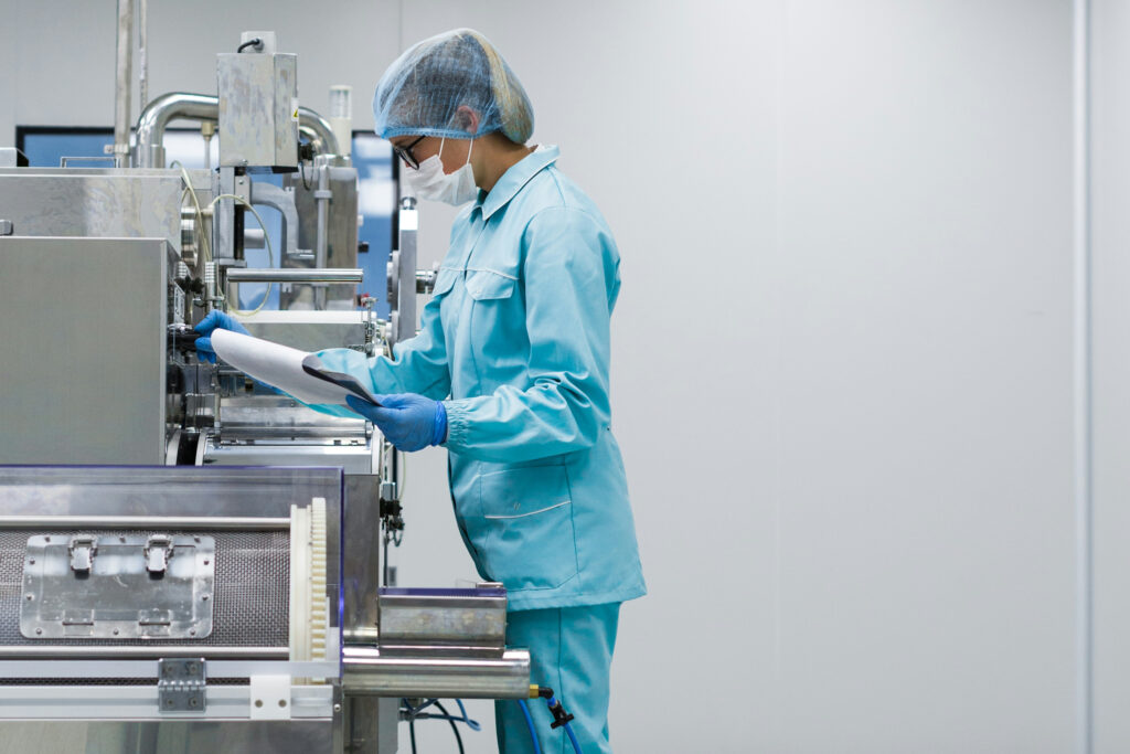 A woman wearing glasses, a face mask, blue PPE and gloves, checks a machine in a pharma manufacturing facility.