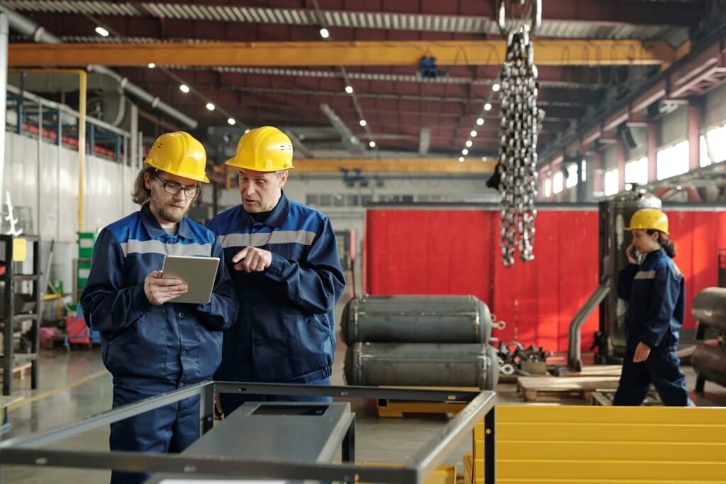 Two men wearing blue overalls and yellow hard hats look at a mobile device whilst standing in front of machinery.