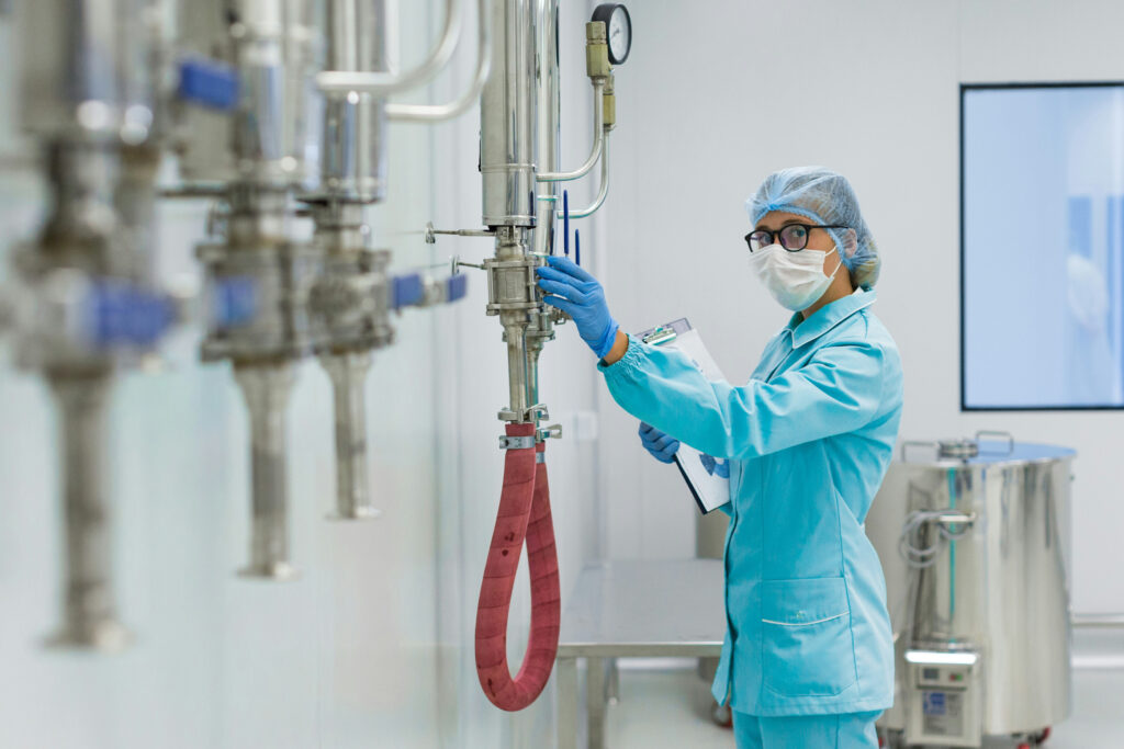 A woman wearing glasses, face mask, blue PPE and gloves adjusts piping in a pharma manufacturing facility