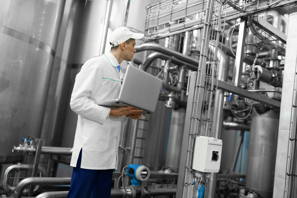 A man wearing a white cap and lab coat looks at piping in a manufacturing facility whilst holding a laptop.