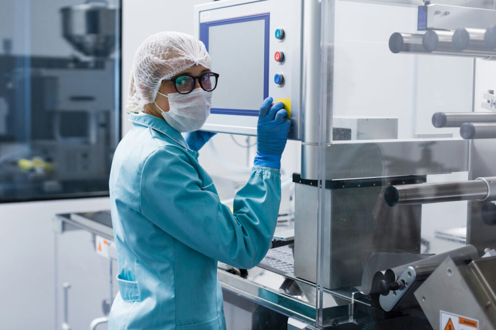 A woman in a white hairnet, face mask and blue lab coat, wearing black glasses and blue latex gloves operates a machine in a clean pharmaceutical factory.