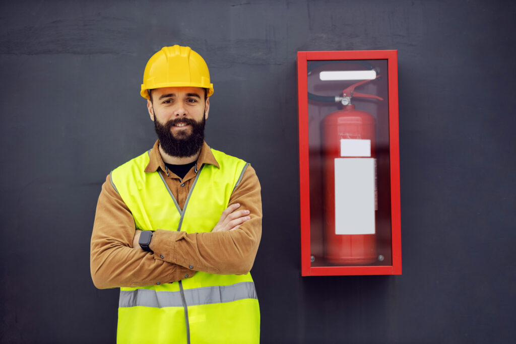 A bearded man in a high-vis vest and yellow hard hat stands beside a wall-mounted fire extinguisher with his arms folded.