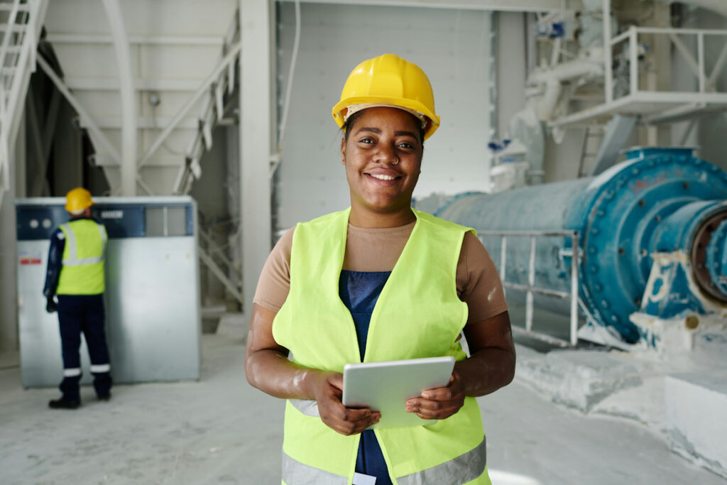 A woman wearing a high-vis vest and yellow hard hat stands in a factory holding a tablet, in the background is a man wearing a high-vis vest and yellow hard hat inspecting some machinery.