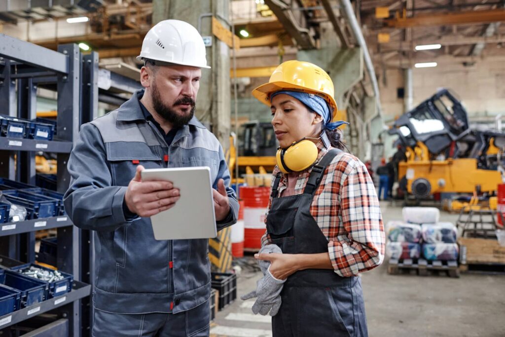 A man in white hardhat holds a tablet and chats with a woman in blue overalls, checked shirt, yellow hard hat and ear defenders.