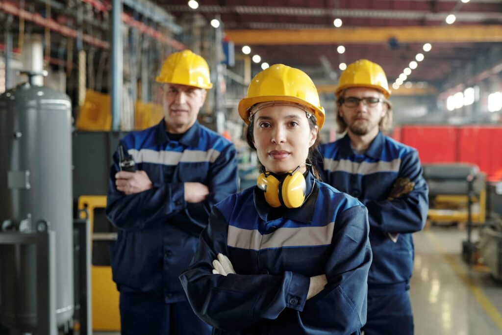 A woman and two men stand with arms folded in a manufacturing facility wearing blue overalls, yellow hard hats and ear defenders.