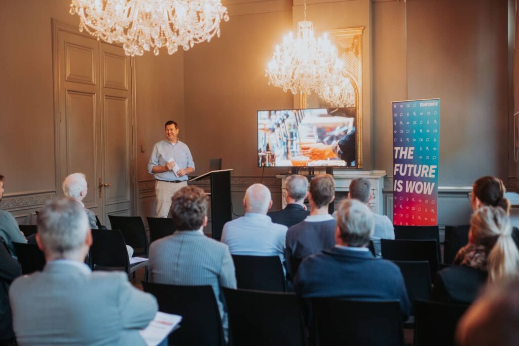 Jan Van den Nieuwenhof, the CEO of TenForce stands at the front of a room presenting beside a large TV screen.