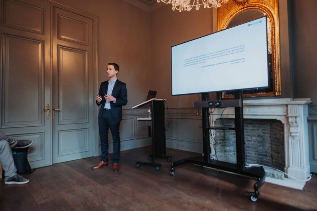 Uros Milosevic from TenForce stands at the front of a meeting room, presenting beside a large TV screen.