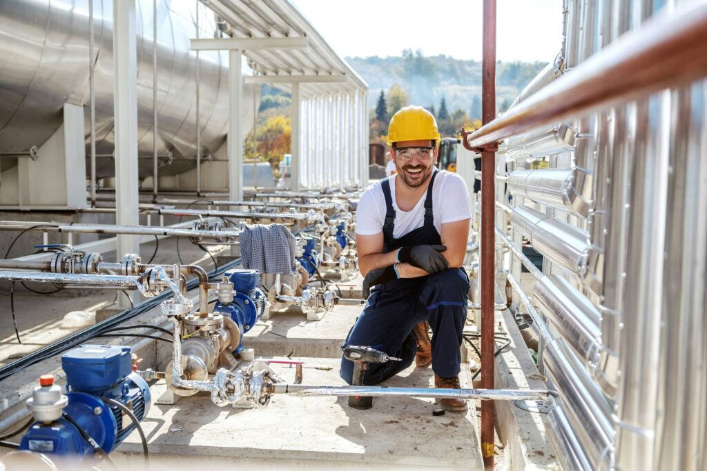 A man wearing a yellow hard hat, safety goggles, gloves and blue overalls crouches, smiling at the camera, amongst silver pipes at a chemical plant.